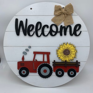 May include: A white wooden round sign with the word "Welcome" in black lettering. A red toy tractor with a black and gray tire is pulling a wagon with a yellow sunflower. A brown burlap bow is at the top of the sign.