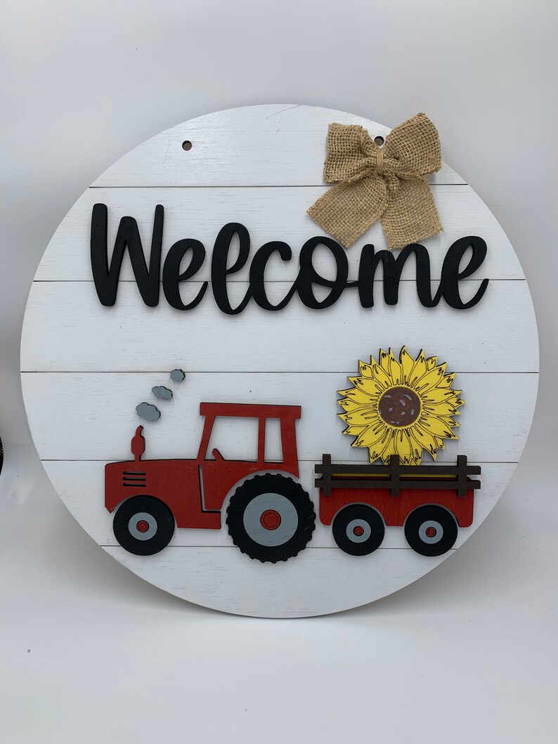 May include: A white wooden round sign with the word "Welcome" in black lettering. A red toy tractor with a black and gray tire is pulling a wagon with a yellow sunflower. A brown burlap bow is at the top of the sign.