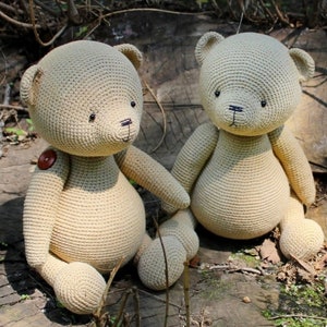 Peut inclure: Deux ours en peluche beige crochet&eacute;s, assis c&ocirc;te &agrave; c&ocirc;te. Les ours ont des yeux en bouton, des nez cousus noirs et un bouton sur l'un d'eux. Ils sont assis sur une surface en bois vieilli, avec un fond de verdure.
