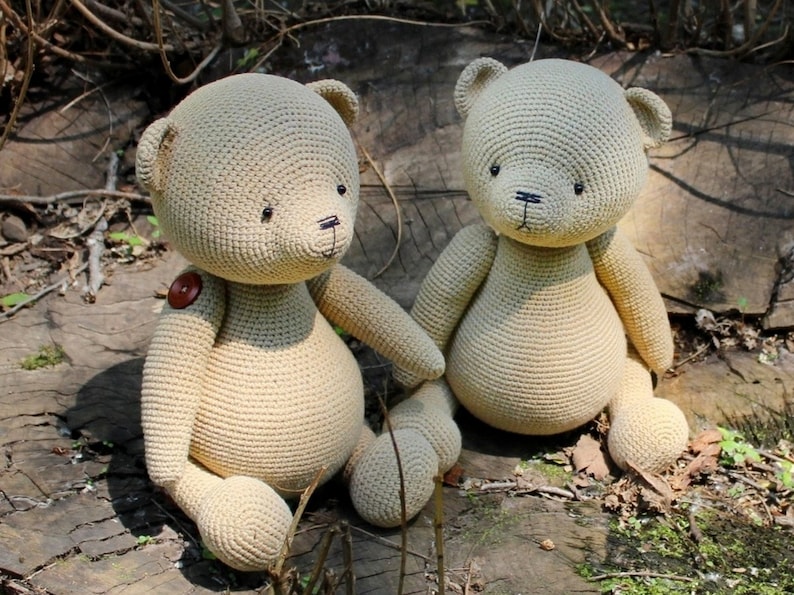 Peut inclure: Deux ours en peluche beige crochet&eacute;s, assis c&ocirc;te &agrave; c&ocirc;te. Les ours ont des yeux en bouton, des nez cousus noirs et un bouton sur l'un d'eux. Ils sont assis sur une surface en bois vieilli, avec un fond de verdure.