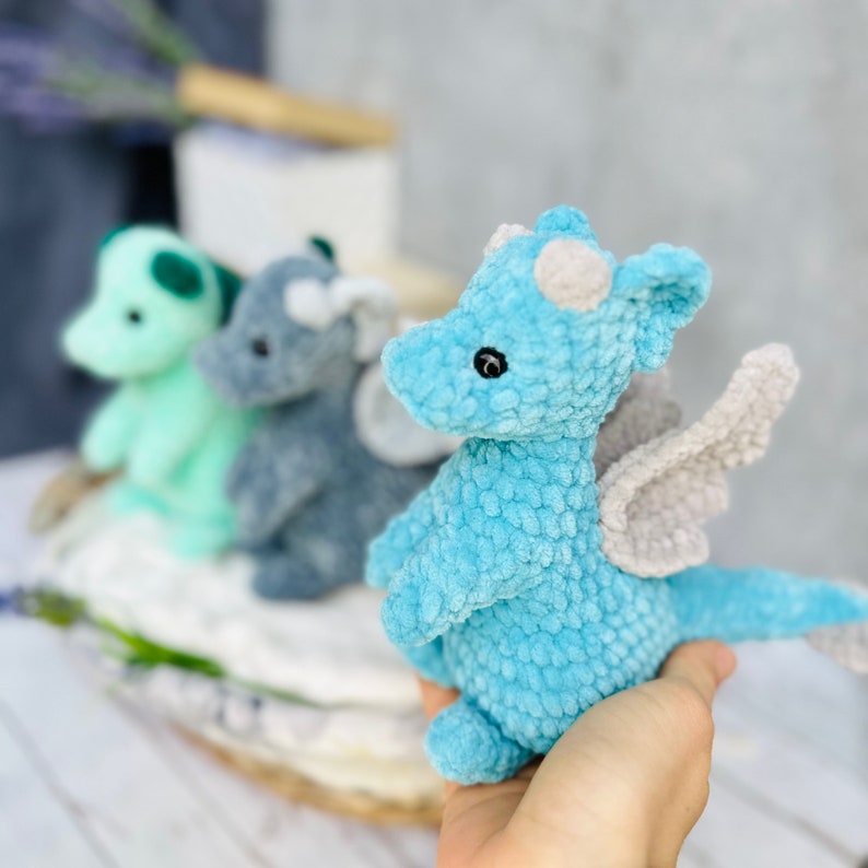 May include: A close-up of three crocheted dragon toys in shades of blue and green. The front-most dragon is light blue with gray wings and white spots. The other two are in the background, one green and one gray. The toys are soft and plush.