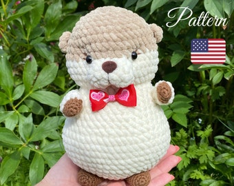 May include: A crocheted otter plush toy with a white and brown body, brown feet, and a red bow with pink hearts. The toy is sitting on a person's hand.