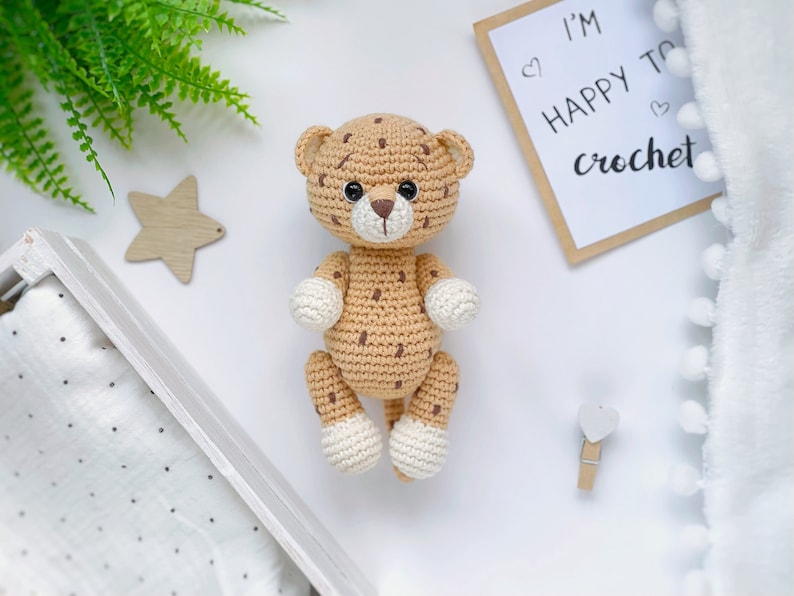 Op de afbeelding: Een handgemaakt gehaakt luipaardspeeltje met een beige lichaam met donkerbruine vlekken, een witte snuit en witte pootjes. Het luipaard heeft zwarte veiligheidsoogjes en een bruin geborduurde neus. Een bordje ernaast toont I'M HAPPY TO crochet met hartjes.