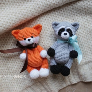 Puede incluir: Dos animales de peluche tejidos a mano, un zorro y un mapache. El zorro naranja tiene patas blancas y una cinta marr&oacute;n. El mapache gris tiene patas negras, un antifaz negro y una cinta azul claro. El texto @KNIT.FRIENDS es visible a la izquierda.