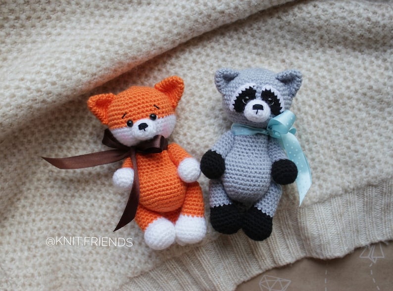 Puede incluir: Dos animales de peluche tejidos a mano, un zorro y un mapache. El zorro naranja tiene patas blancas y una cinta marr&oacute;n. El mapache gris tiene patas negras, un antifaz negro y una cinta azul claro. El texto @KNIT.FRIENDS es visible a la izquierda.
