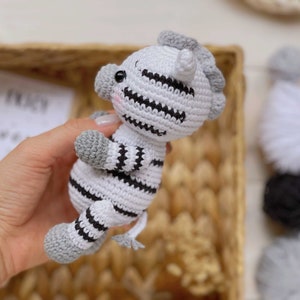 May include: A small, handmade crochet zebra toy featuring a white body with black horizontal stripes, grey hooves, a grey mane, and a small grey tail. The plush animal has black safety eyes and subtle pink blush detailing on its cheeks.