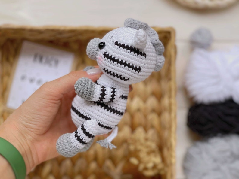 May include: A small, handmade crochet zebra toy featuring a white body with black horizontal stripes, grey hooves, a grey mane, and a small grey tail. The plush animal has black safety eyes and subtle pink blush detailing on its cheeks.