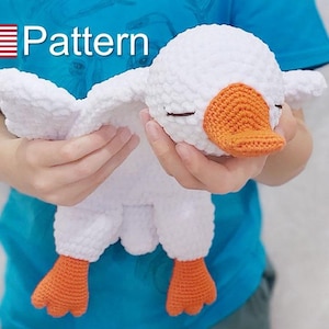 Op de afbeelding: Een wit gebreid knuffelgoosje met oranje pootjes en een oranje snavel. De gans heeft zijn ogen dicht en wordt vastgehouden door een persoon.
