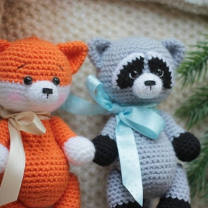 Puede incluir: Dos animales de peluche tejidos a ganchillo a mano, un zorro y un mapache. El zorro es naranja y blanco con una cinta beige. El mapache es gris, negro y blanco con una cinta azul claro de lunares. Ambos tienen ojos de pl&aacute;stico negro.