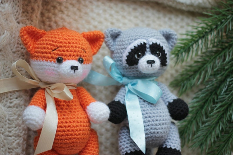 Puede incluir: Dos animales de peluche tejidos a ganchillo a mano, un zorro y un mapache. El zorro es naranja y blanco con una cinta beige. El mapache es gris, negro y blanco con una cinta azul claro de lunares. Ambos tienen ojos de pl&aacute;stico negro.