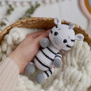 May include: A small, handcrafted crochet zebra toy featuring a white body with black horizontal stripes, grey hooves, and a grey snout. The zebra has black safety eyes, small ears, and subtle pink blush detailing on its cheeks, created with yarn.