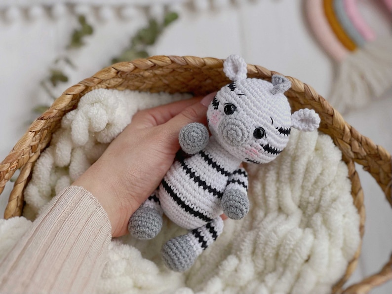 May include: A small, handcrafted crochet zebra toy featuring a white body with black horizontal stripes, grey hooves, and a grey snout. The zebra has black safety eyes, small ears, and subtle pink blush detailing on its cheeks, created with yarn.