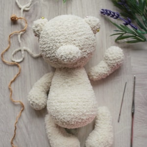 May include: A white crocheted teddy bear with a fluffy texture. The bear is laying on a light wood surface with yarn and needles nearby.