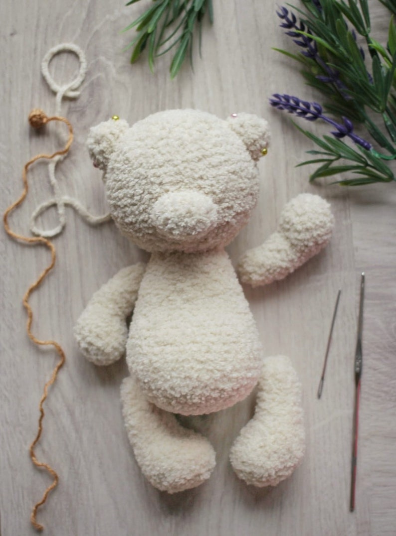 May include: A white crocheted teddy bear with a fluffy texture. The bear is laying on a light wood surface with yarn and needles nearby.