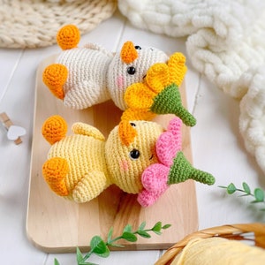May include: Two crocheted ducklings with orange feet and beaks, black eyes, and pink blush marks. One duckling is cream-colored with a yellow flower hat, and the other is yellow with a pink flower hat, both featuring green stems on top.
