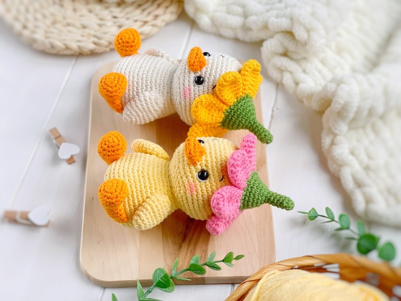 May include: Two crocheted ducklings with orange feet and beaks, black eyes, and pink blush marks. One duckling is cream-colored with a yellow flower hat, and the other is yellow with a pink flower hat, both featuring green stems on top.