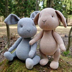 Op de afbeelding: Twee gehaakte konijnenknuffels, &eacute;&eacute;n lichtgrijs en &eacute;&eacute;n lichtbruin, zittend op een mossige boomstam in een bosrijke omgeving.