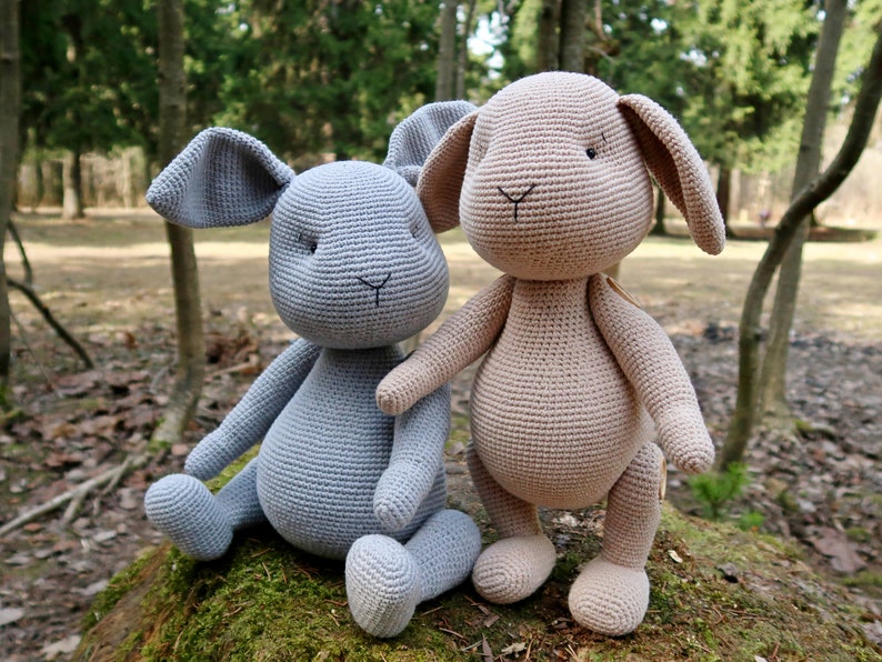 Op de afbeelding: Twee gehaakte konijnenknuffels, &eacute;&eacute;n lichtgrijs en &eacute;&eacute;n lichtbruin, zittend op een mossige boomstam in een bosrijke omgeving.