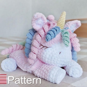 Puede incluir: Un juguete de unicornio de crochet con una melena rosa, azul y blanca y un cuerno amarillo. El unicornio está acostado con los ojos cerrados. El juguete está sobre una superficie blanca y marrón. Patrón.