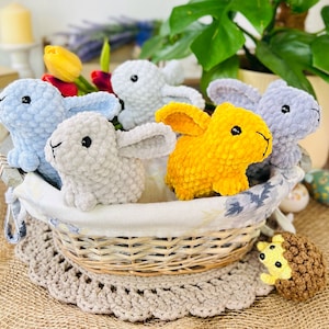 Puede incluir: Una cesta de mimbre llena de juguetes de conejo de crochet. Los conejos son de varios colores, incluyendo azul claro, gris y amarillo. La cesta est&aacute; sentada sobre una superficie de arpillera marr&oacute;n.