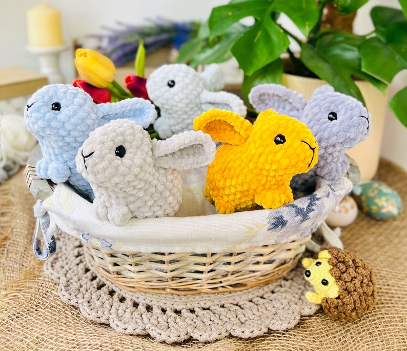 Puede incluir: Una cesta de mimbre llena de juguetes de conejo de crochet. Los conejos son de varios colores, incluyendo azul claro, gris y amarillo. La cesta est&aacute; sentada sobre una superficie de arpillera marr&oacute;n.