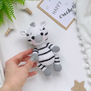 May include: A small, handmade crochet zebra toy featuring a white body with black stripes, grey hooves, and a grey snout. The zebra has black safety eyes, embroidered eyebrows, and soft pink blush on its cheeks. It is a stuffed, soft-textured animal.