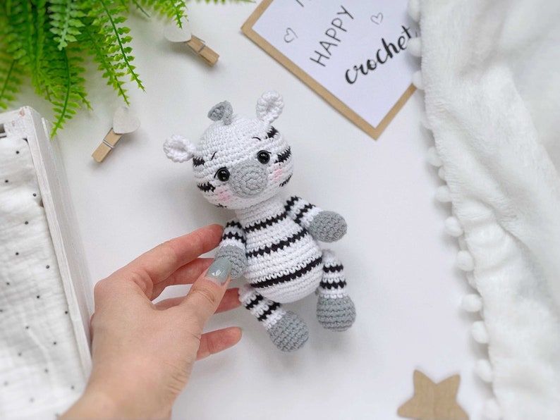 May include: A small, handmade crochet zebra toy featuring a white body with black stripes, grey hooves, and a grey snout. The zebra has black safety eyes, embroidered eyebrows, and soft pink blush on its cheeks. It is a stuffed, soft-textured animal.