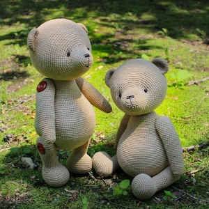 Peut inclure: Deux ours en peluche au crochet beiges sont pr&eacute;sent&eacute;s en ext&eacute;rieur. L'un est debout avec des boutons, l'autre est assis. Les ours ont des yeux et un nez noirs. L'arri&egrave;re-plan est de l'herbe verte et du feuillage.