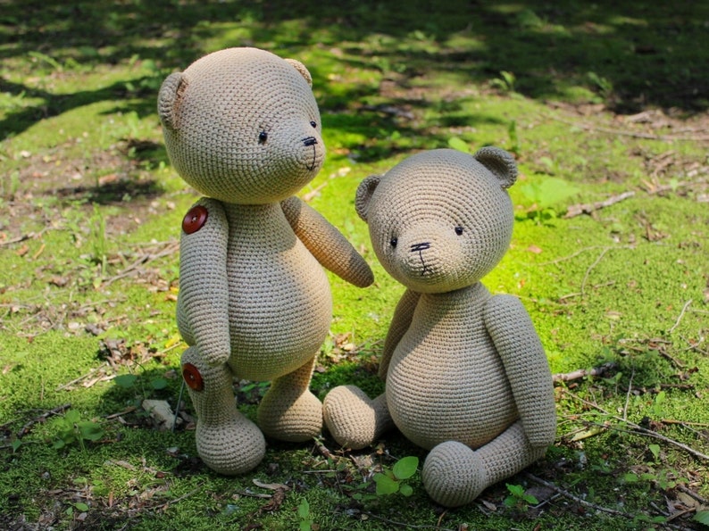 Peut inclure: Deux ours en peluche au crochet beiges sont pr&eacute;sent&eacute;s en ext&eacute;rieur. L'un est debout avec des boutons, l'autre est assis. Les ours ont des yeux et un nez noirs. L'arri&egrave;re-plan est de l'herbe verte et du feuillage.