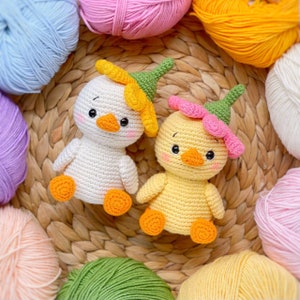 May include: Two crocheted ducklings, one white and one yellow, each wearing a green pointed hat topped with a flower. The white duckling has a yellow flower, and the yellow duckling has a pink flower. Both have orange beaks and feet.