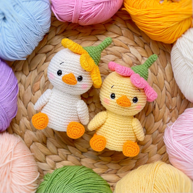 May include: Two crocheted ducklings, one white and one yellow, each wearing a green pointed hat topped with a flower. The white duckling has a yellow flower, and the yellow duckling has a pink flower. Both have orange beaks and feet.