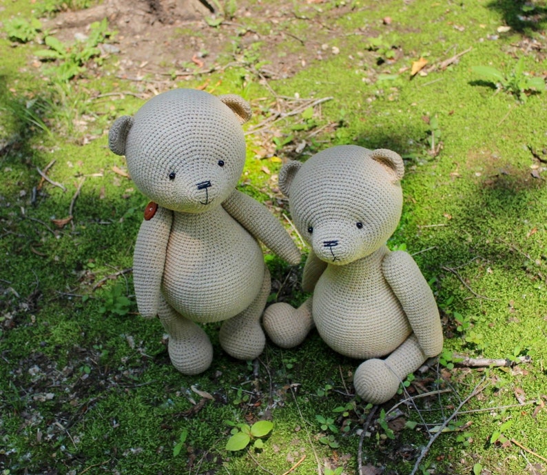 Peut inclure: Deux ours en peluche beige crochet&eacute;s sont assis sur de l'herbe verte. L'un se tient debout avec un bouton sur la poitrine, tandis que l'autre est assis. Les deux ont des yeux noirs et des traits cousus. Les ours sont faits &agrave; la main.