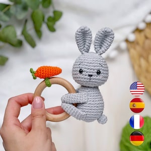 Pode incluir: Um brinquedo de coelho de crochê cinza com uma cenoura presa a um anel de dentição de madeira. O coelho tem olhos pretos e um pequeno nariz rosa.