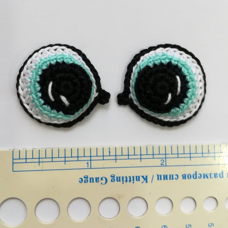 May include: Two crocheted eyes with black pupils, white scleras, and turquoise irises. The eyes are approximately 1 inch in diameter.
