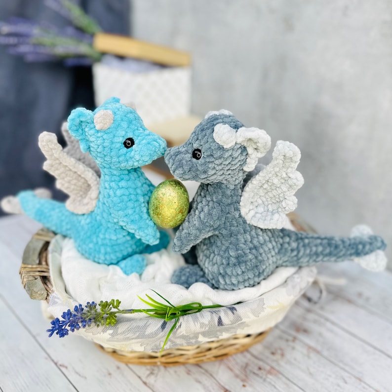 May include: Two handmade crochet dragons, one light blue and one gray, with white and gray wings, sit in a woven basket. They are facing each other, with a gold egg between them. The dragons have small horns and are made of soft yarn.