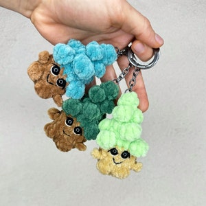 May include: Three crocheted keychains shaped like trees. The trees are green, blue, and brown. Each tree has a smiling face.