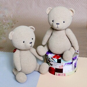 Peut inclure: Deux ours en peluche beige crochet&eacute;s. L'un est assis sur une bo&icirc;te ronde d&eacute;corative avec des motifs floraux et textuels. L'autre est assis sur une surface bleu clair. Les deux ours ont des yeux noirs et des bouches cousues.