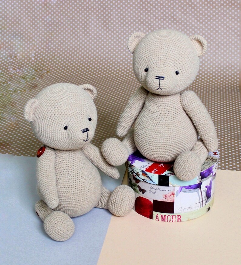 Peut inclure: Deux ours en peluche beige crochet&eacute;s. L'un est assis sur une bo&icirc;te ronde d&eacute;corative avec des motifs floraux et textuels. L'autre est assis sur une surface bleu clair. Les deux ours ont des yeux noirs et des bouches cousues.