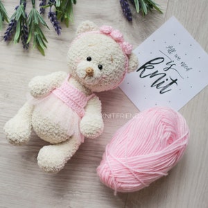 May include: A white crocheted teddy bear wearing a pink crocheted dress and a pink bow. The bear is lying on a wooden surface with a ball of pink yarn and a sign that says "All we need is knit".