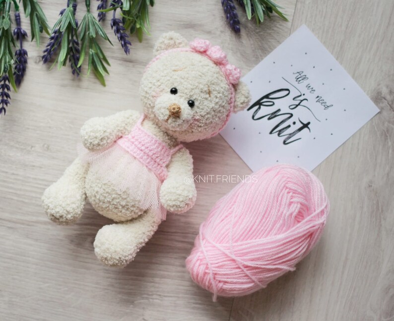 May include: A white crocheted teddy bear wearing a pink crocheted dress and a pink bow. The bear is lying on a wooden surface with a ball of pink yarn and a sign that says "All we need is knit".