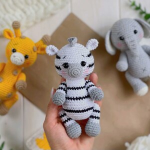 May include: A collection of three handmade crochet stuffed animals including a yellow giraffe with brown spots, a white and black striped zebra, and a grey elephant. Each animal features embroidered facial details, pink cheeks, and rounded limbs.
