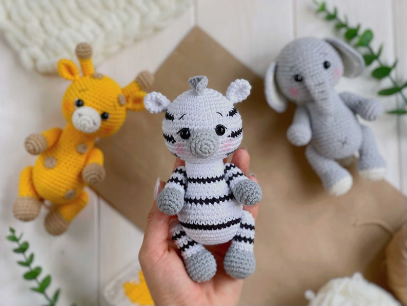 May include: A collection of three handmade crochet stuffed animals including a yellow giraffe with brown spots, a white and black striped zebra, and a grey elephant. Each animal features embroidered facial details, pink cheeks, and rounded limbs.