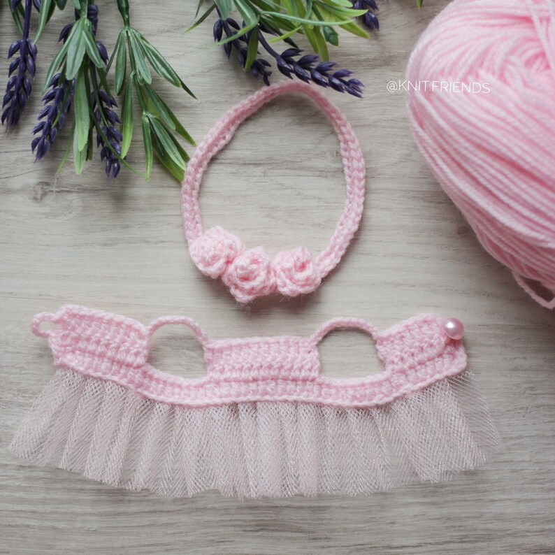 May include: A pink crocheted headband with three crocheted flower embellishments and a pink crocheted dress with a tulle skirt and a pearl bead.