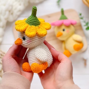 May include: A cream-colored crocheted duckling toy wearing a bright yellow flower hat with a green center. The duckling has an orange beak and feet, and small black eyes with pink blush accents. A second yellow crocheted duckling with a pink flower hat is visible.