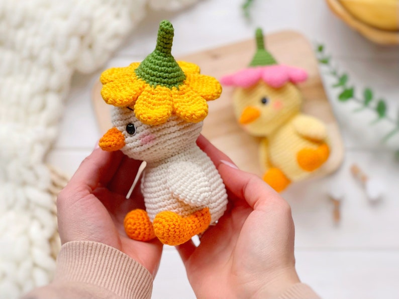 May include: A cream-colored crocheted duckling toy wearing a bright yellow flower hat with a green center. The duckling has an orange beak and feet, and small black eyes with pink blush accents. A second yellow crocheted duckling with a pink flower hat is visible.