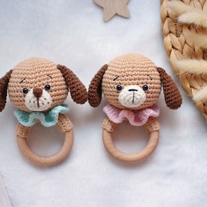 Puede incluir: Dos juguetes de sonajero de perro de crochet con anillos de madera. Un perro tiene un collar azul claro y el otro un collar rosa.