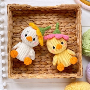 May include: Two small crocheted ducklings with round bodies and orange feet. One duckling is white with a yellow flower hat, and the other is yellow with a pink flower hat. Both have black eyes, orange beaks, and pink blush on their cheeks.