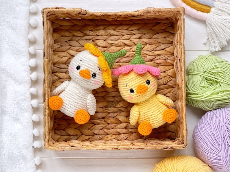 May include: Two small crocheted ducklings with round bodies and orange feet. One duckling is white with a yellow flower hat, and the other is yellow with a pink flower hat. Both have black eyes, orange beaks, and pink blush on their cheeks.