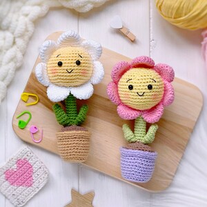 May include: Two crocheted flower pot decorations. One flower is white with a yellow center and the other is pink with a yellow center. Both flowers have smiling faces.