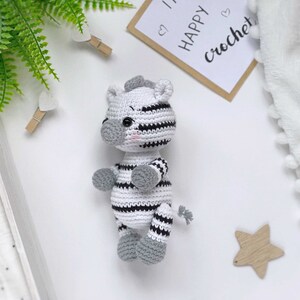 May include: A small, handmade crochet zebra toy featuring a white body with black horizontal stripes, a grey snout, grey ears, grey feet, and a small grey tail. The zebra has black safety eyes and subtle pink blush details on its cheeks.